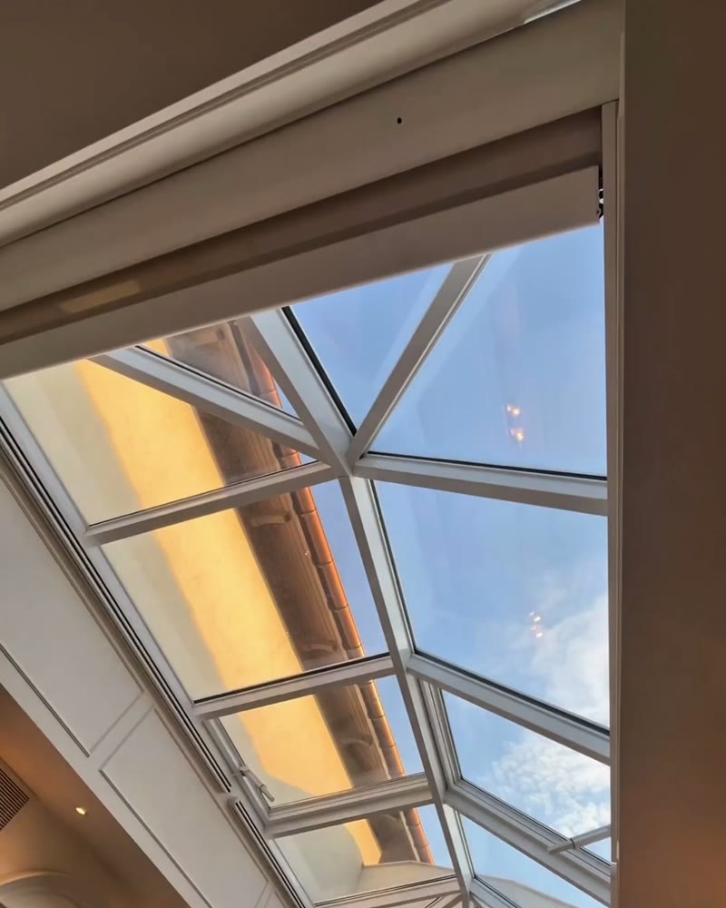 Fabric ceiling shade partly open under a glass roof inside a house