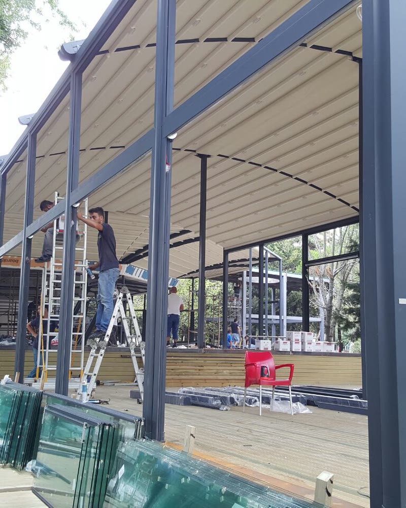 Installers fitting a retractable fabric roof and glass walls over a restaurant terrace among trees