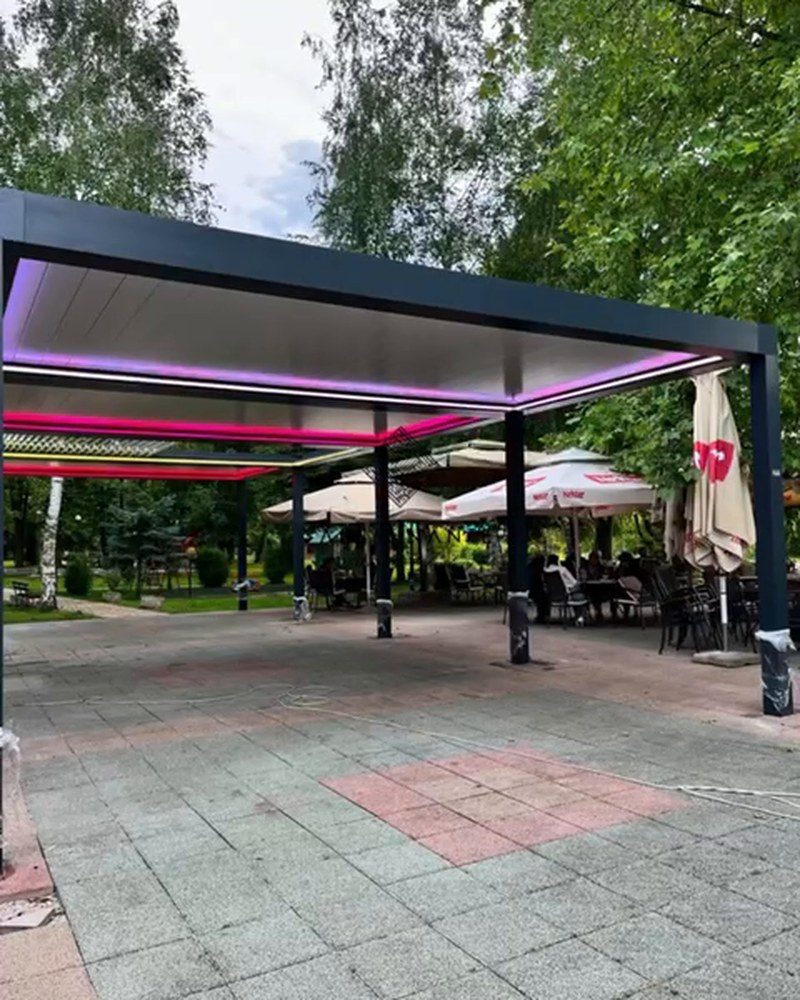Louvered pergolas with colored LED lighting over a paved restaurant terrace among trees