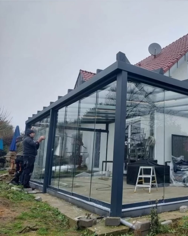 Glass room with a dark aluminum frame being finished by installers next to a house