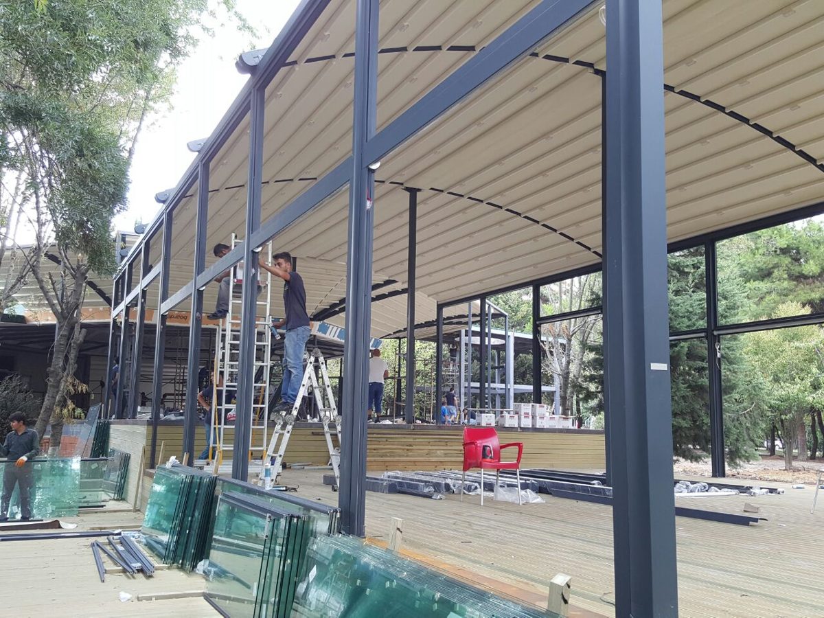 Installers on ladders fitting glass walls under a retractable fabric roof on a restaurant terrace